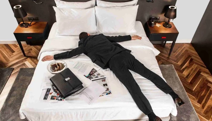 Businessman in a suit lying exhausted on a hotel bed with documents and a briefcase, showcasing the comfort of Cue Hotel rooms.