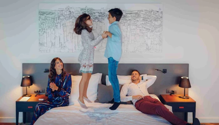 Family enjoying quality time on a large bed; two children jumping while parents watch and smile in a modern hotel room.