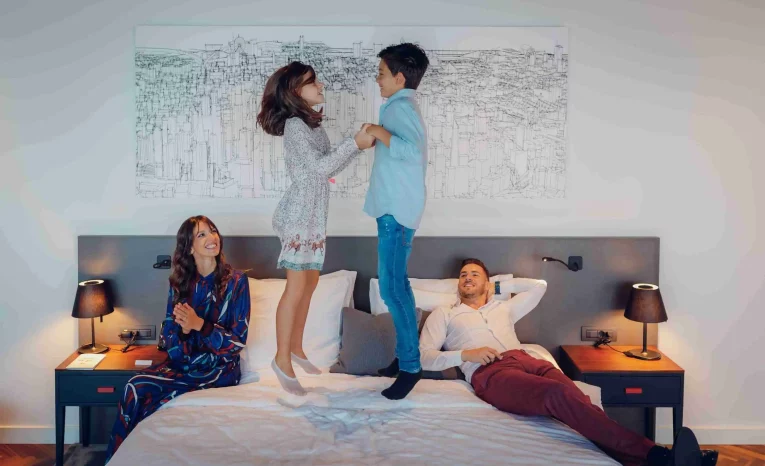 Family enjoying quality time on a large bed; two children jumping while parents watch and smile in a modern hotel room.